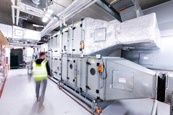 Mitie male employee, blurred and wearing a hi vis vest, walking in a room containing large manufacturing equipment